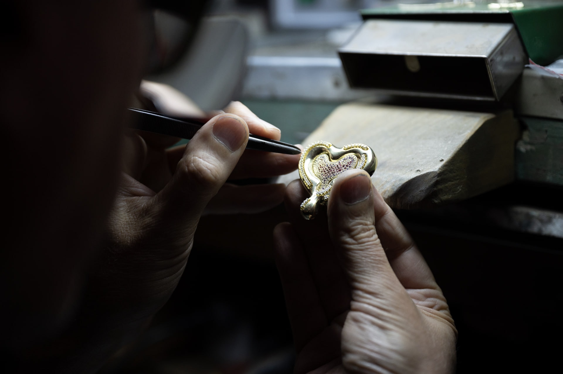 A goldsmith at the bench finishing a gold and gemstone heart pendant with tweezers
