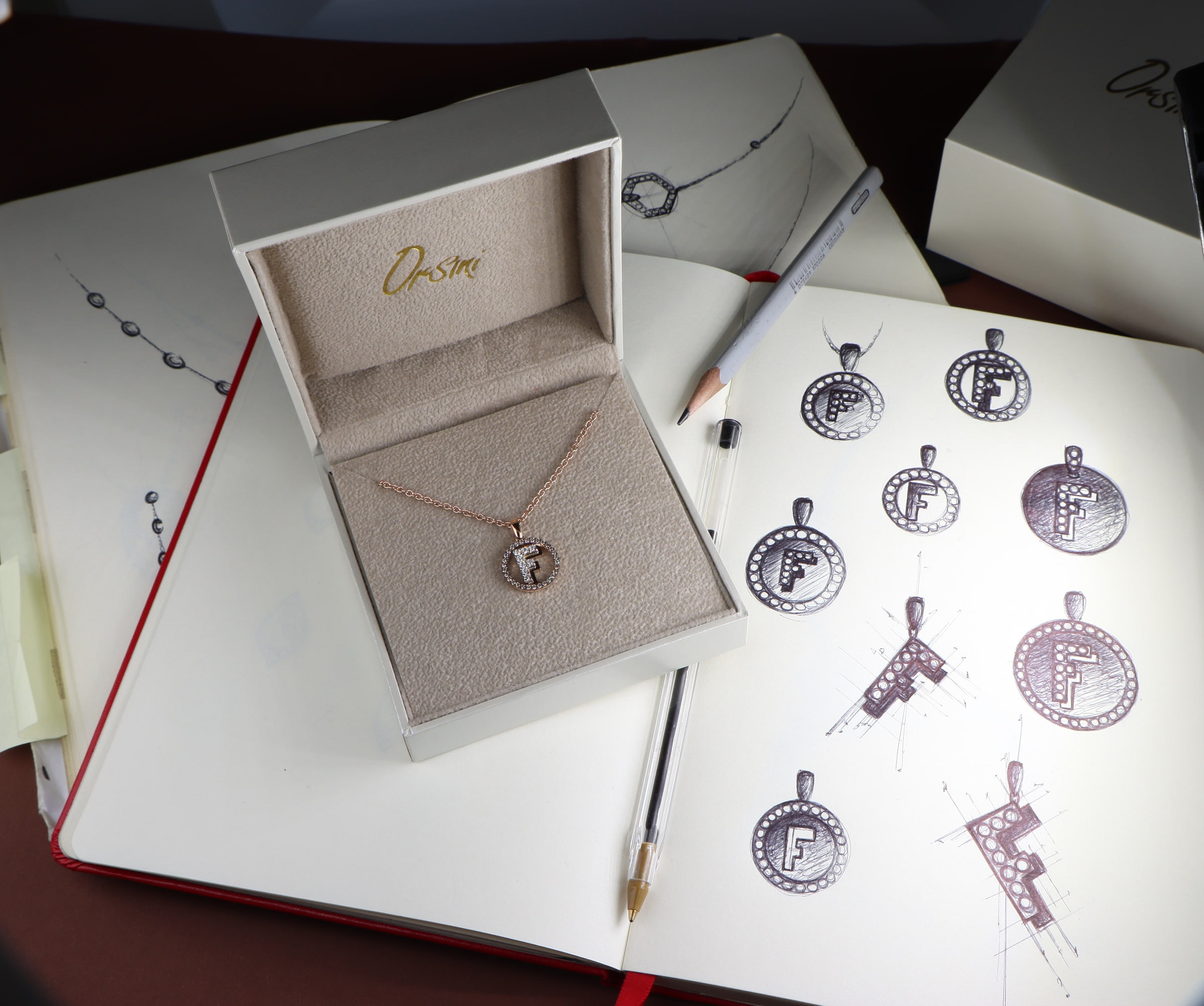A pendant with the initial F in its Orsini box, beside the sketchbook it came from